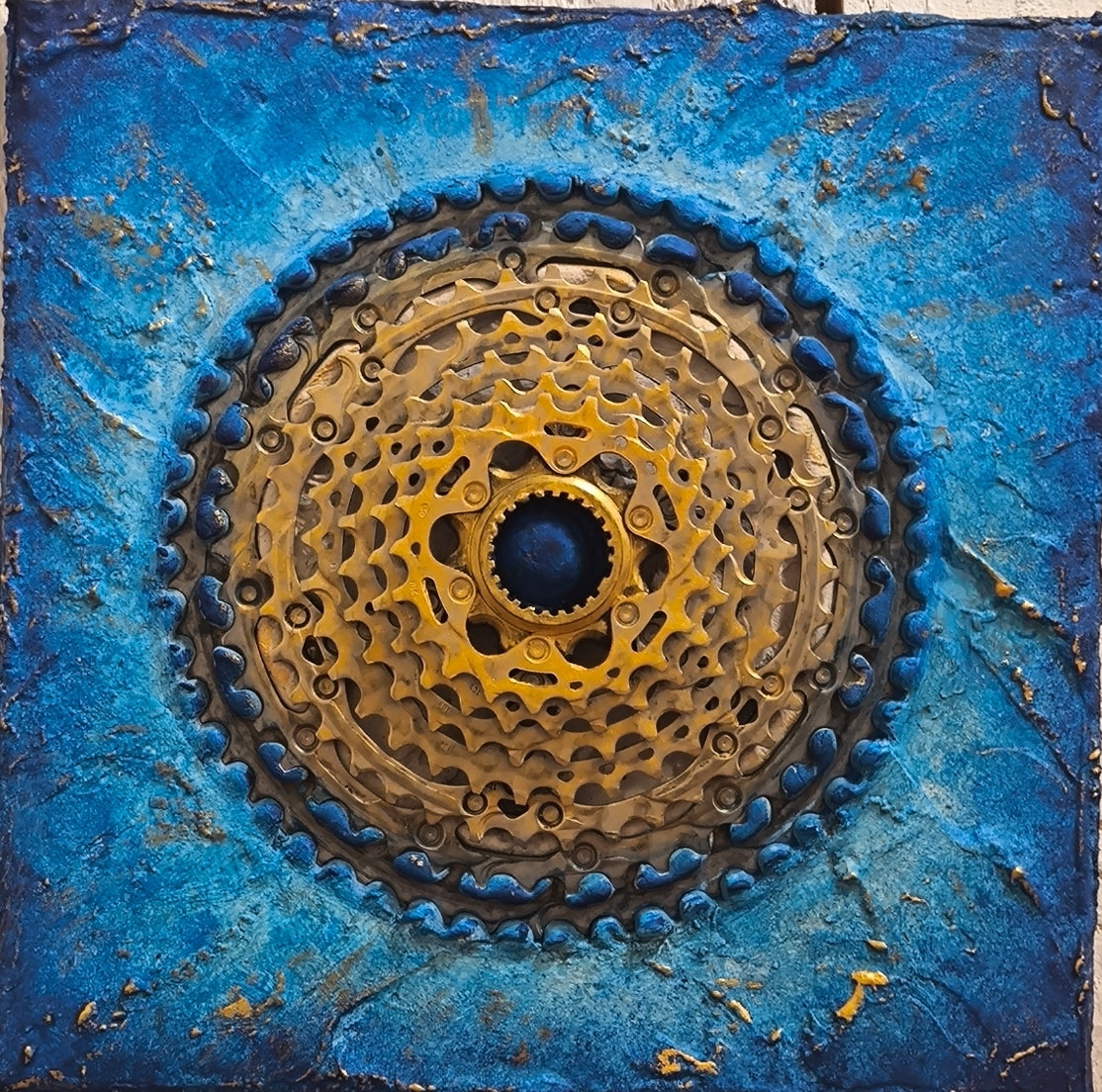 Dans cette œuvre, François Tapiézo célèbre le mouvement perpétuel à travers un assemblage unique de pièces industrielles et de cycle. La cassette de vélo, élément central de la composition, forme une pyramide dorée qui symbolise la dynamique solaire et l'énergie infinie du mouvement.
Le contraste entre les matériaux bruts – acier, bois, sables – et la douceur des pigments de Provence rehaussés à l'or crée une harmonie visuelle captivante. L'or appliqué au pinceau de soie sublime chaque détail des textures.